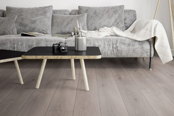 Wohnzimmer mit stilvollem Laminatboden, der echtem Holz täuschen ähnlich sieht. Auf dem Boden steht eine helle Couch und davor ein kleiner Couchtisch.